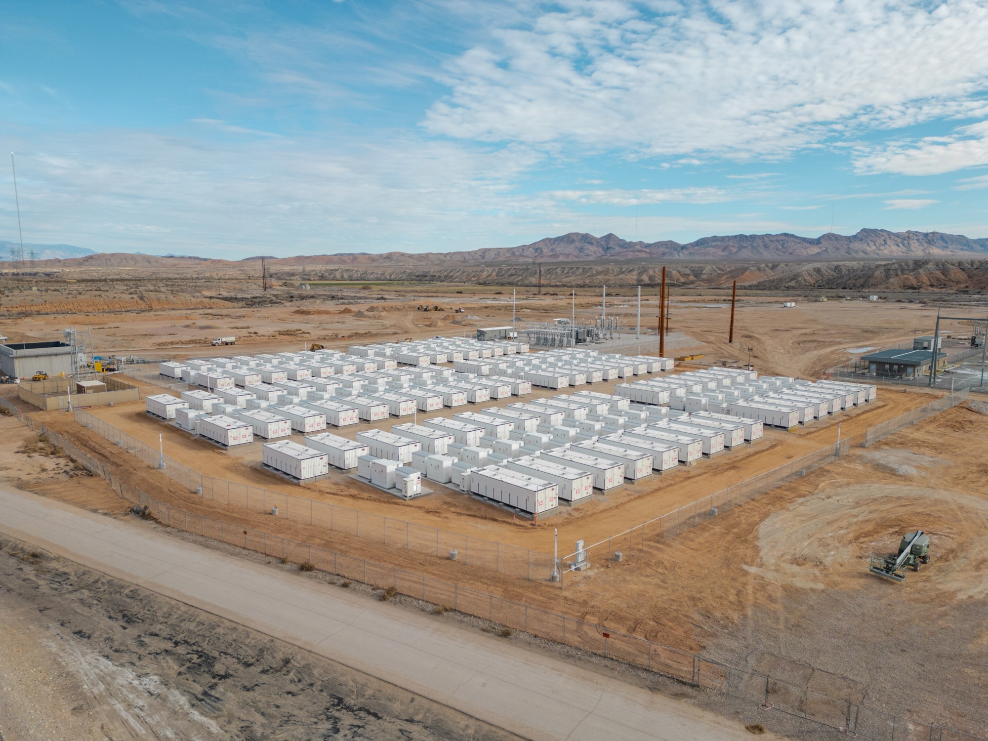 Aerial view of rows of white battery enclosures behind a fence at a utility-scale storage site in the desert.